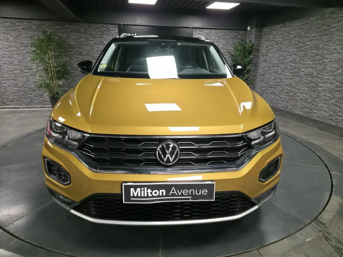 Vue avant d'un Volkswagen T-Roc jaune Curcuma avec calandre noire et détails chromés en intérieur showroom.