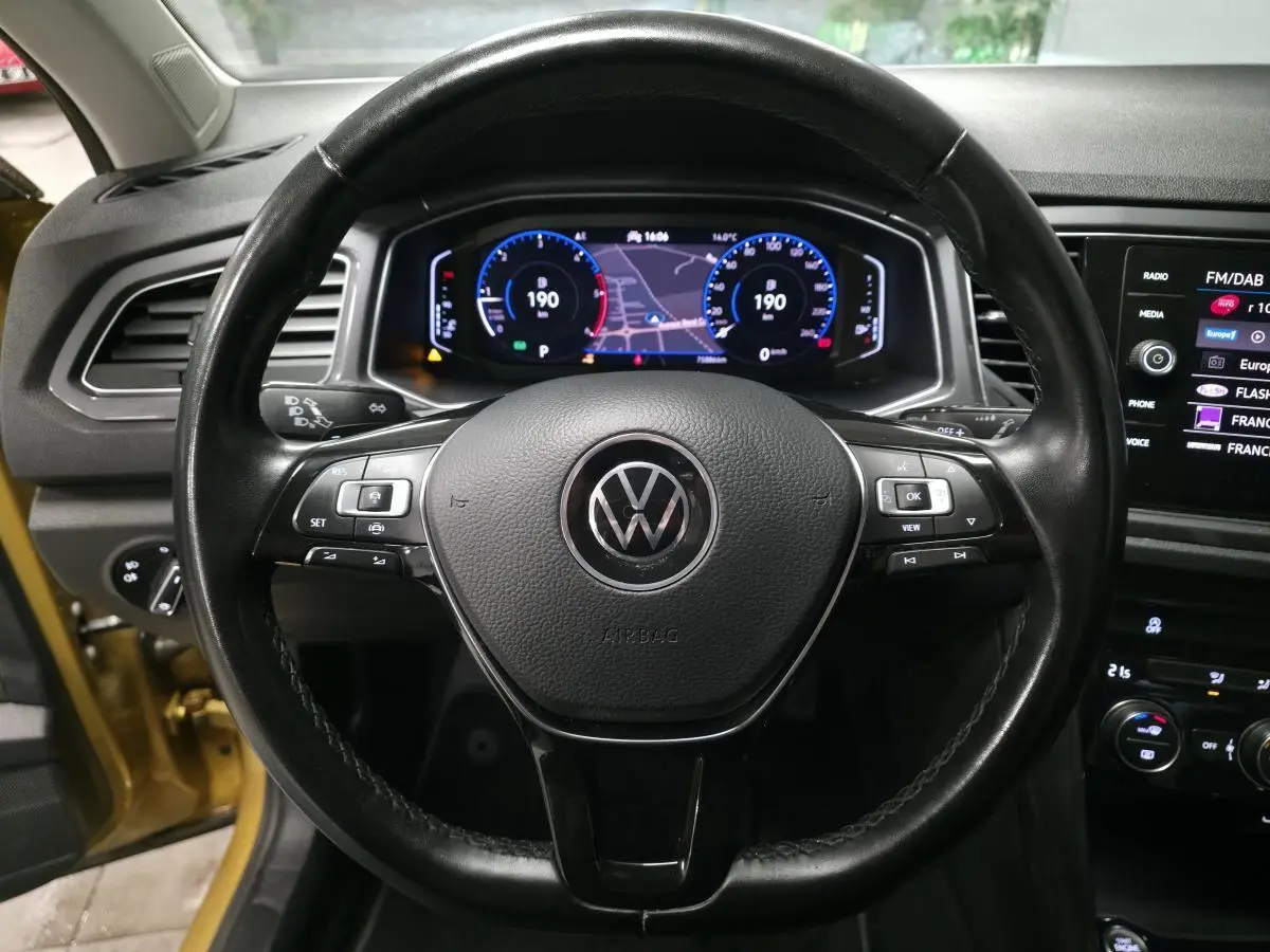 Vue centrée sur le volant en cuir noir d’un Volkswagen T-Roc jaune, avec tableau de bord numérique et écran tactile visible.