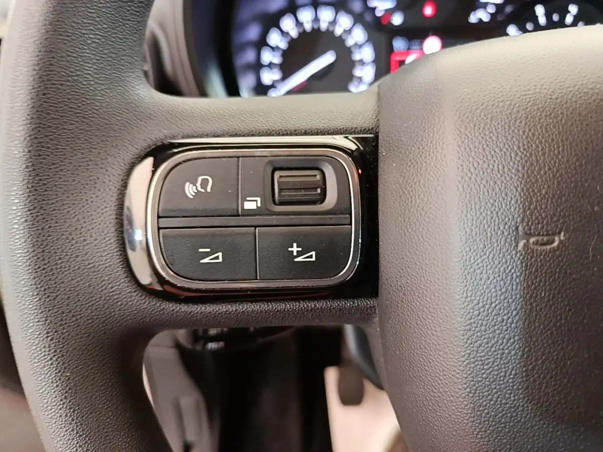 Gros plan sur les commandes multifonctions noires à gauche du volant en cuir noir du Citroën Berlingo blanc, tableau de bord flou en arrière-plan.