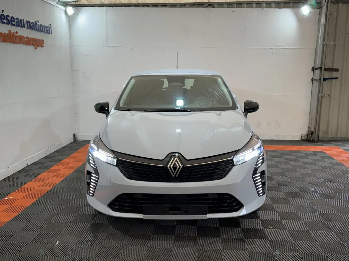 Vue avant d'une Renault Clio gris clair avec phares LED allumés dans un garage au sol noir et orange.