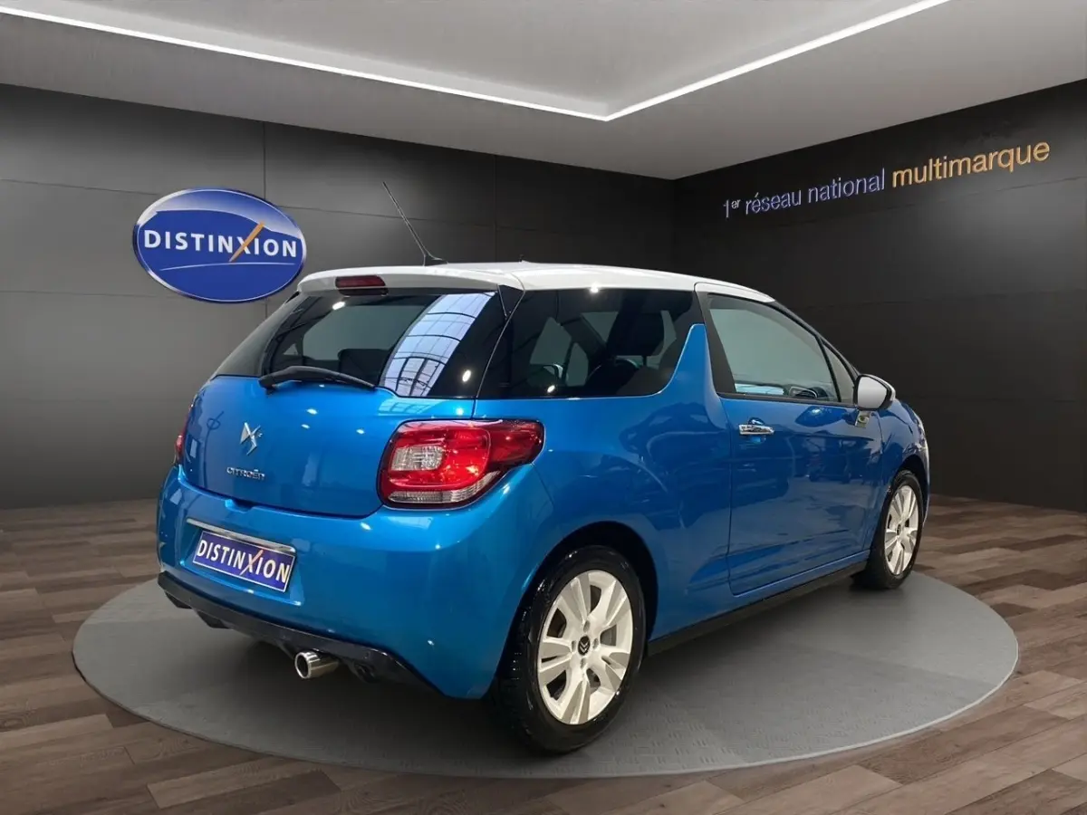 Vue 3/4 arrière droite d'une Citroën DS3 e-HDi 90 Airdream So Chic bleu avec toit blanc en showroom.