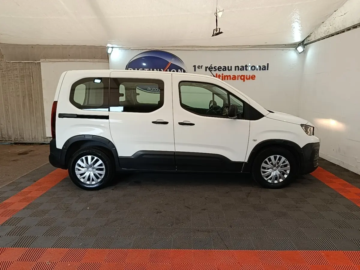 Vue de profil côté gauche d'un Peugeot Rifter blanc 2020 avec protections noires et jantes argentées en intérieur showroom.