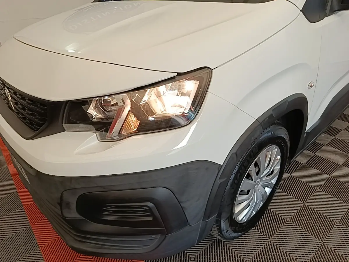 Vue 3/4 avant droit du Peugeot Rifter blanc avec phares allumés et pare-chocs noir mat contrastant.