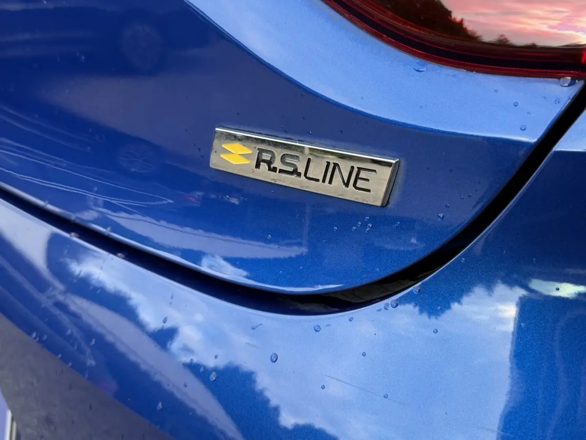 Gros plan sur l'arrière bleu métallisé d'une Renault Clio RS Line avec le badge RS Line visible sous la pluie.