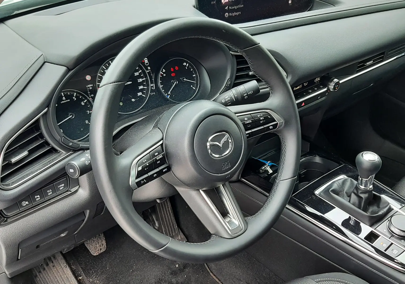 Vue intérieure côté conducteur de la Mazda CX-30 2024, volant cuir, écran central, boîte manuelle et tableau de bord noir.