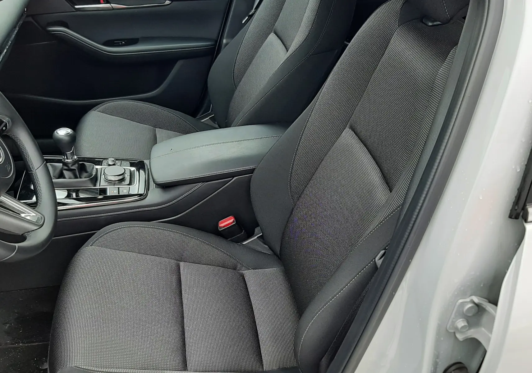 Vue intérieure côté conducteur de la Mazda CX-30 blanche 2024, sièges tissu noir et levier de vitesses manuel visible.