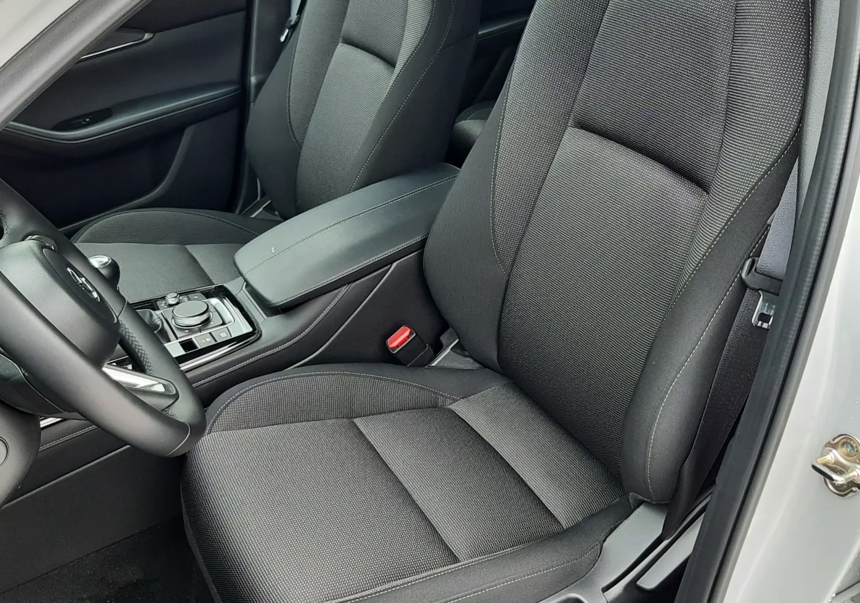 Vue intérieure côté conducteur du Mazda CX-30 2024 avec sièges tissu noir et accoudoir central, portière blanche ouverte.