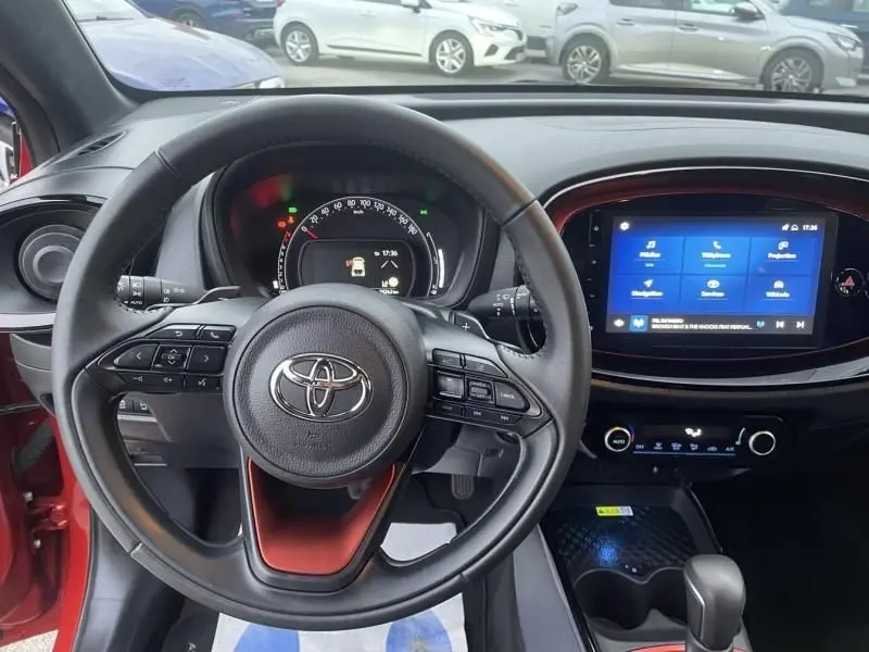 Vue intérieure centrée sur le volant de la Toyota Aygo X rouge piment avec tableau de bord numérique et écran tactile.