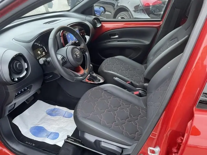 Vue intérieure côté conducteur de la Toyota Aygo X rouge piment avec sellerie noire à motifs et tableau de bord moderne.