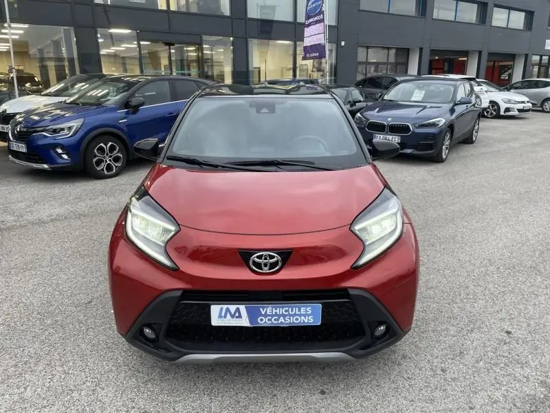 Vue de face d'une Toyota Aygo X 2023 rouge piment avec toit noir, phare allumé, sur parking extérieur.