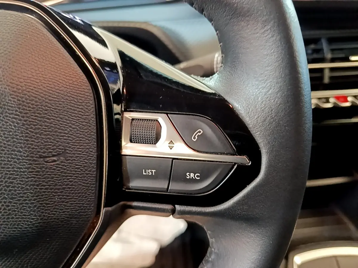 Gros plan sur les commandes gauche du volant cuir noir du Peugeot 2008 blanc, avec boutons multifonctions et molette.