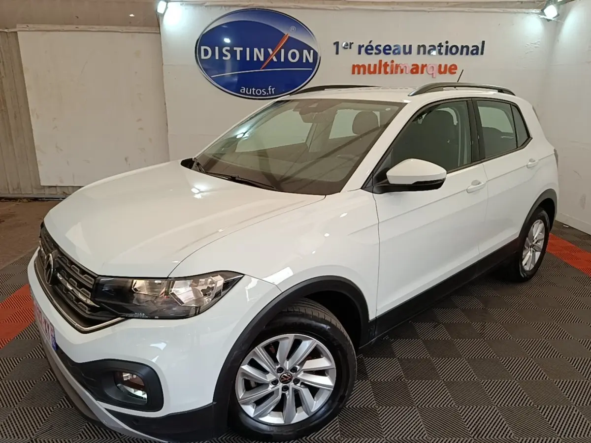 Volkswagen T-Cross blanc vue 3/4 avant droit, avec jantes alliage et barres de toit noires visibles en intérieur showroom.