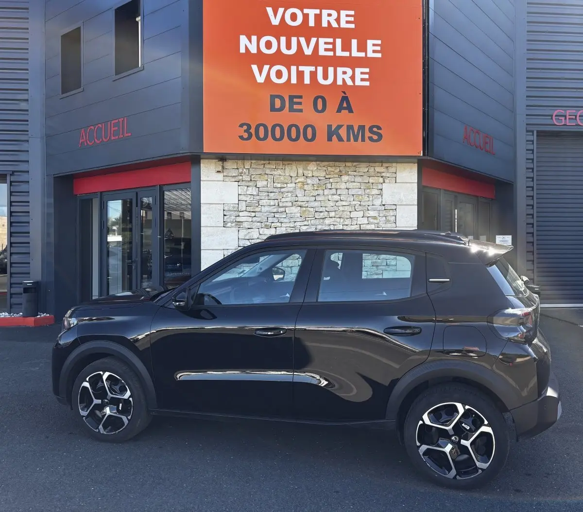 Profil côté gauche d'une Citroën C3 Turbo 100 BVM6 MAX noire, avec jantes distinctives, devant un bâtiment commercial.