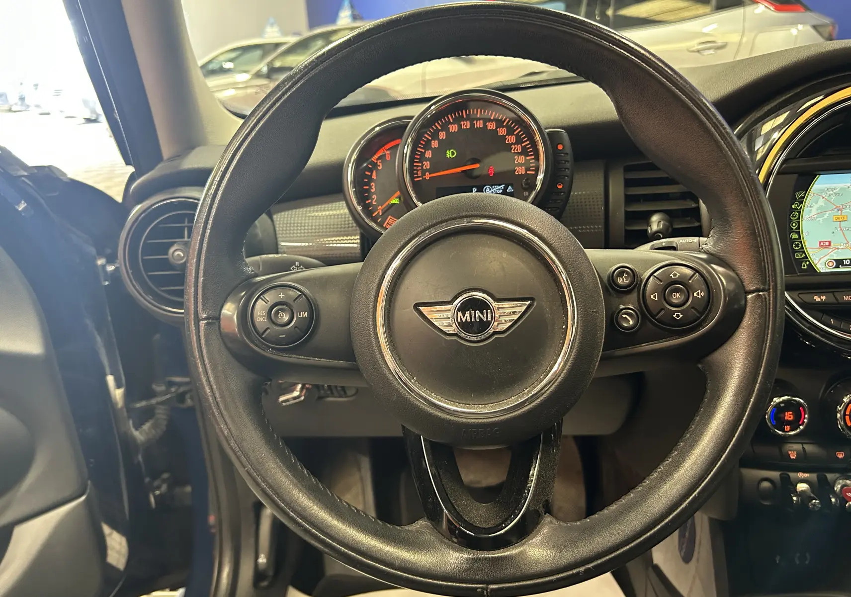 Vue intérieure centrée sur le volant en cuir du MINI 5P Cooper D bleu, avec tableau de bord et écran GPS visibles à droite.