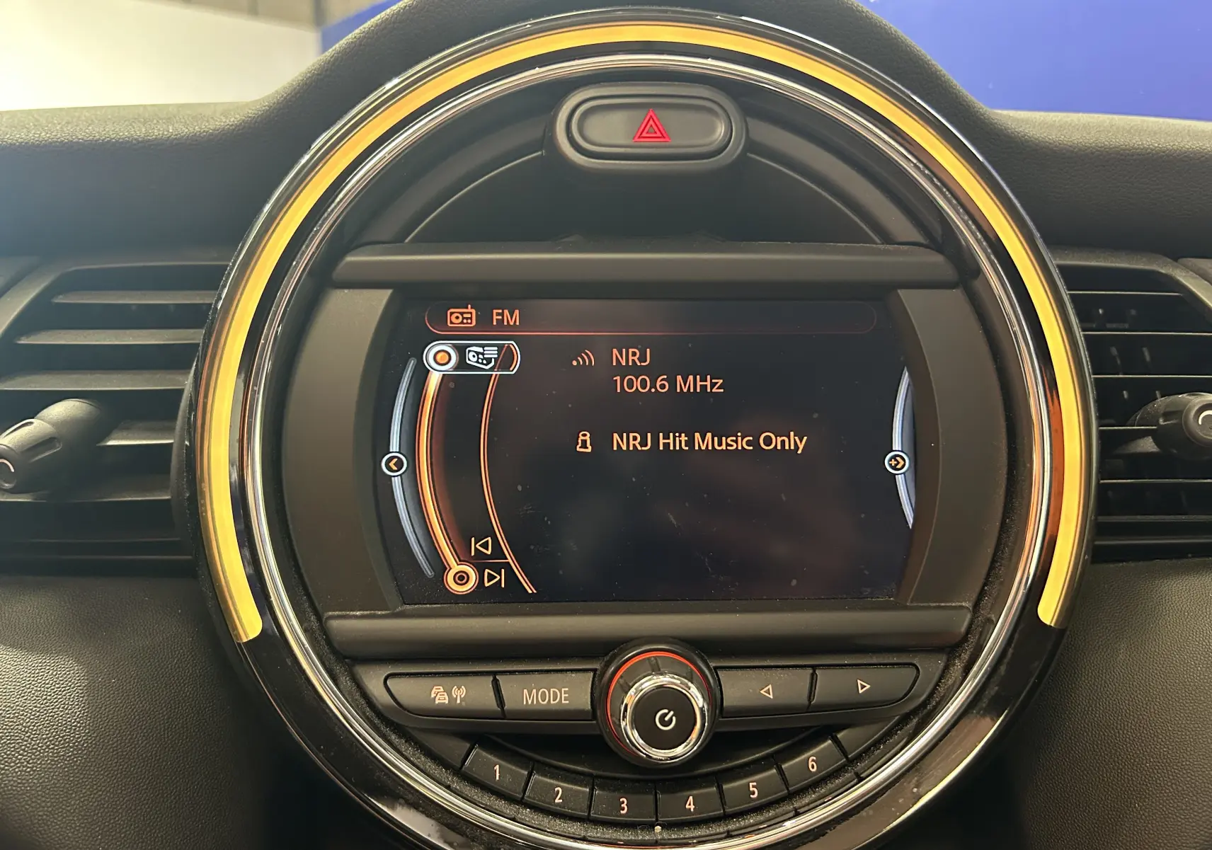 Gros plan sur l’écran central rond du tableau de bord de la MINI 5P bleu, affichant la radio NRJ 100.6 MHz.