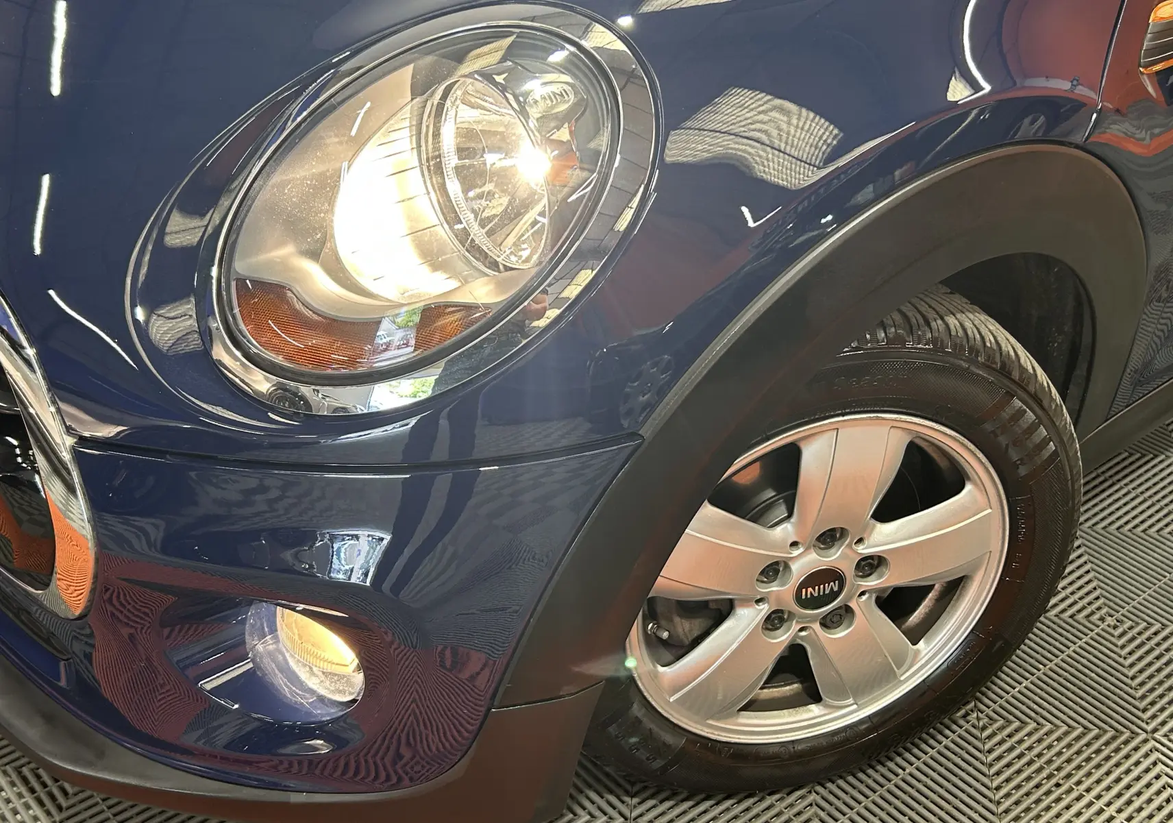 Gros plan sur l'avant droit bleu d'une MINI 5 portes avec phare allumé et jante alu siglée MINI.