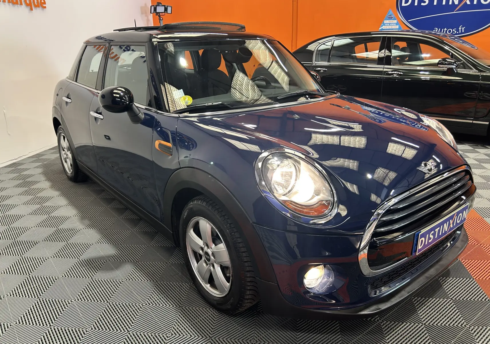 Vue 3/4 avant droite d'une MINI 5 portes Cooper D bleu foncé avec toit ouvrant, phares allumés en intérieur showroom.
