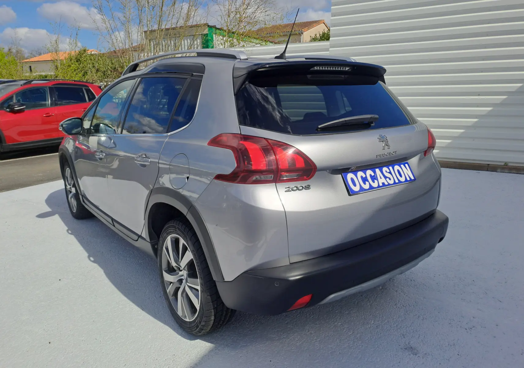 Peugeot 2008 gris vue 3/4 arrière droit, avec barres de toit et feux arrière distinctifs, sur parking extérieur.