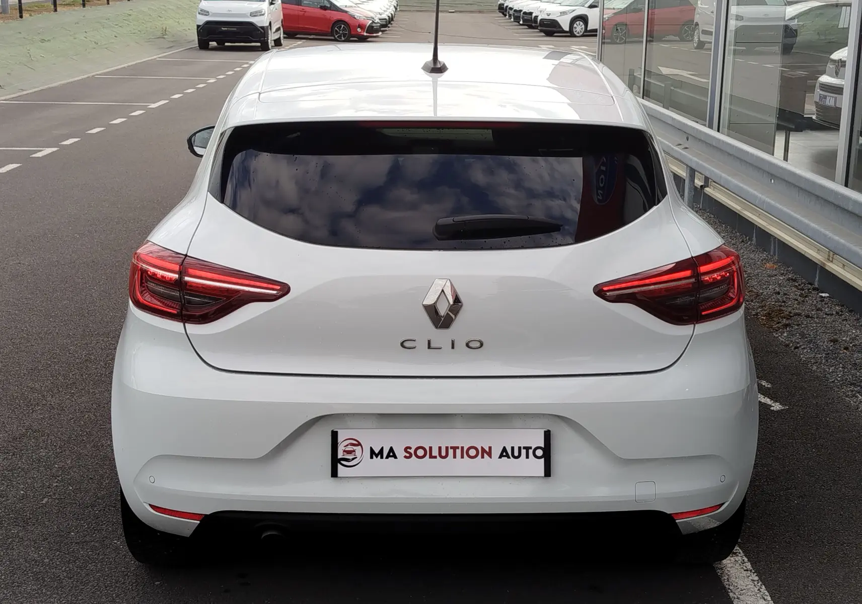 Vue arrière d'une Renault Clio blanche Glacier 2023 avec feux LED et vitre arrière teintée, stationnée devant un bâtiment vitré.
