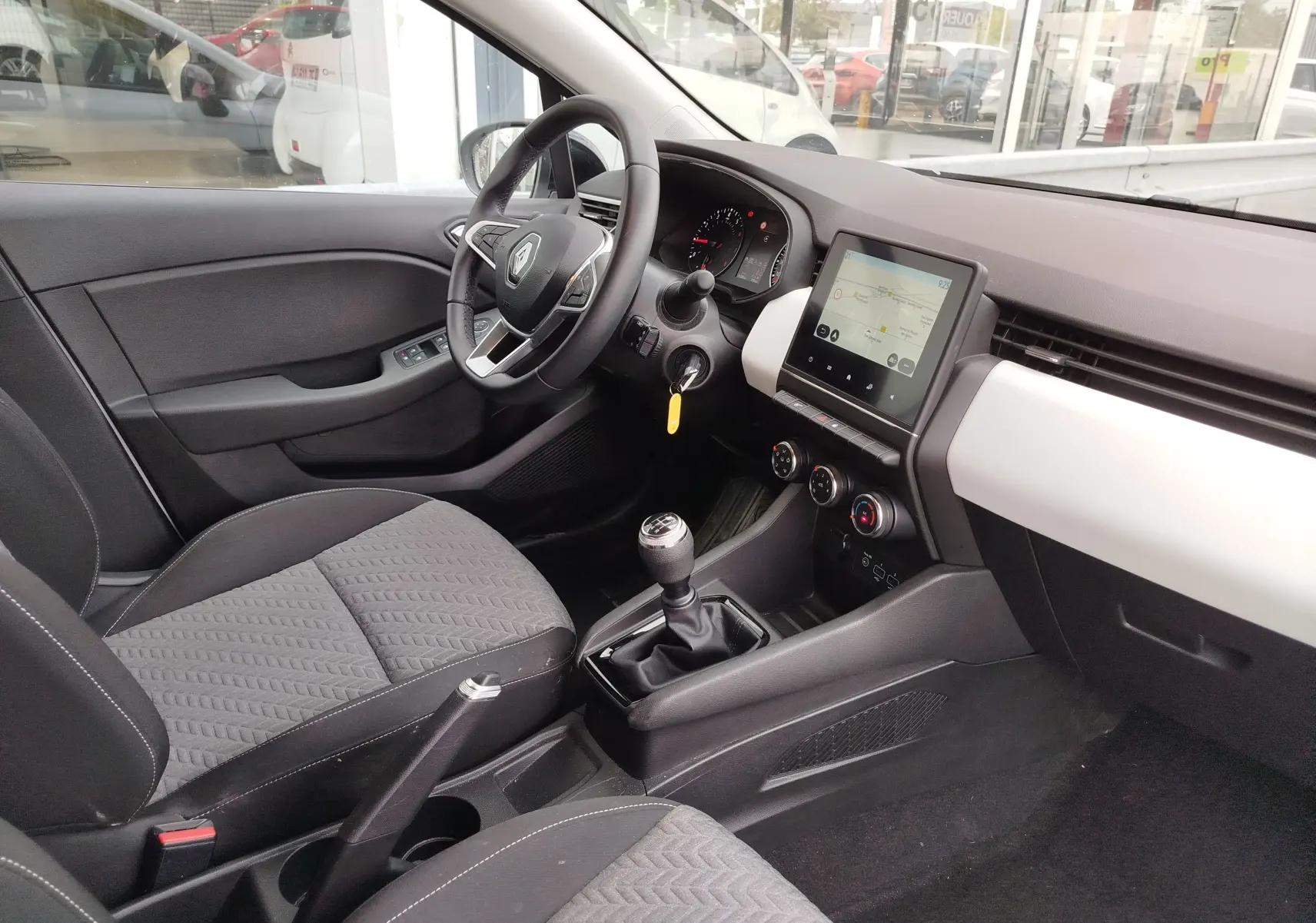 Intérieur de Renault Clio 2023 vu côté conducteur, tableau de bord gris clair avec écran tactile et boîte manuelle.