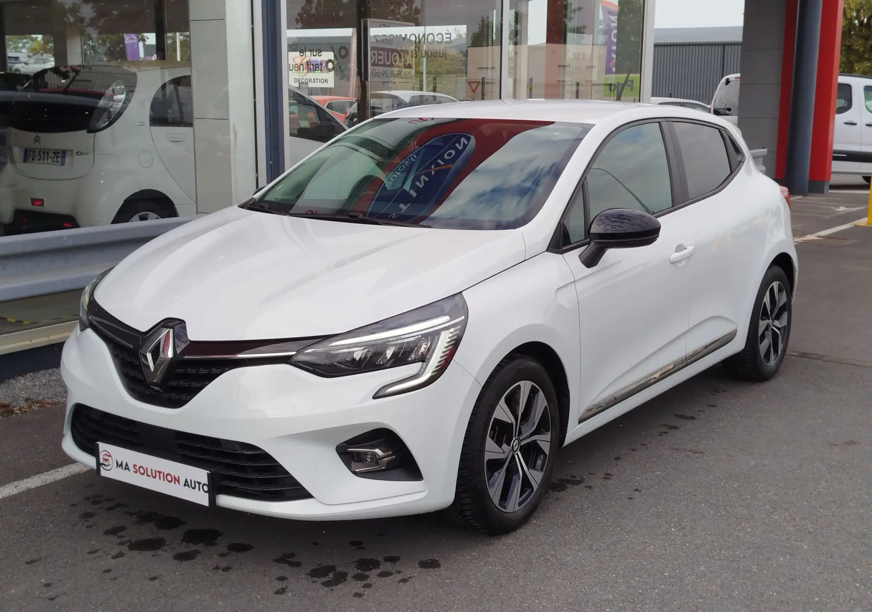 Renault Clio blanc glacier vue 3/4 avant droit, avec jantes alliage bicolores et rétroviseurs noirs.