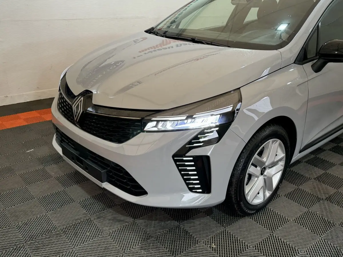 Vue 3/4 avant droit d'une Renault Clio gris clair 2025 avec feux LED et calandre noire distinctive.