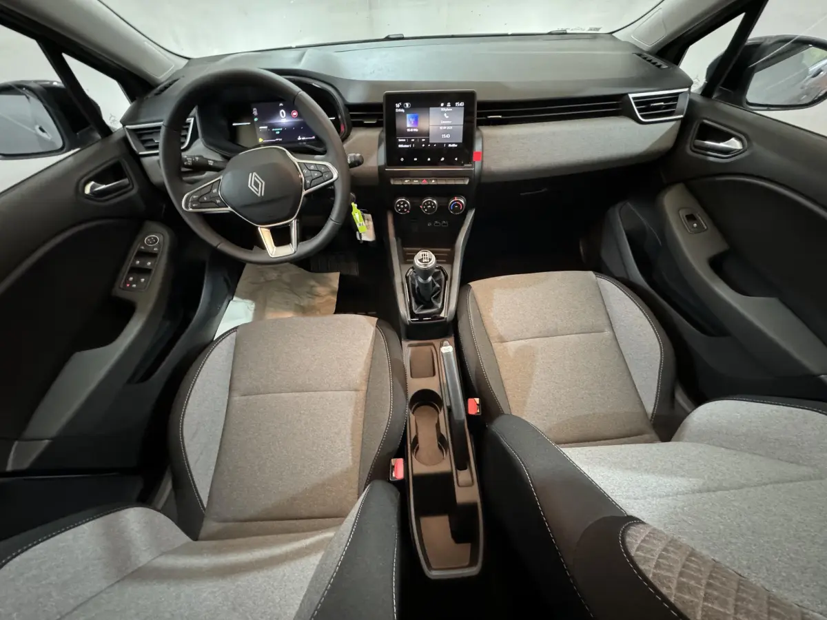 Vue intérieure avant de la Renault Clio gris clair 2025, volant cuir, écran tactile central et boîte manuelle visible.