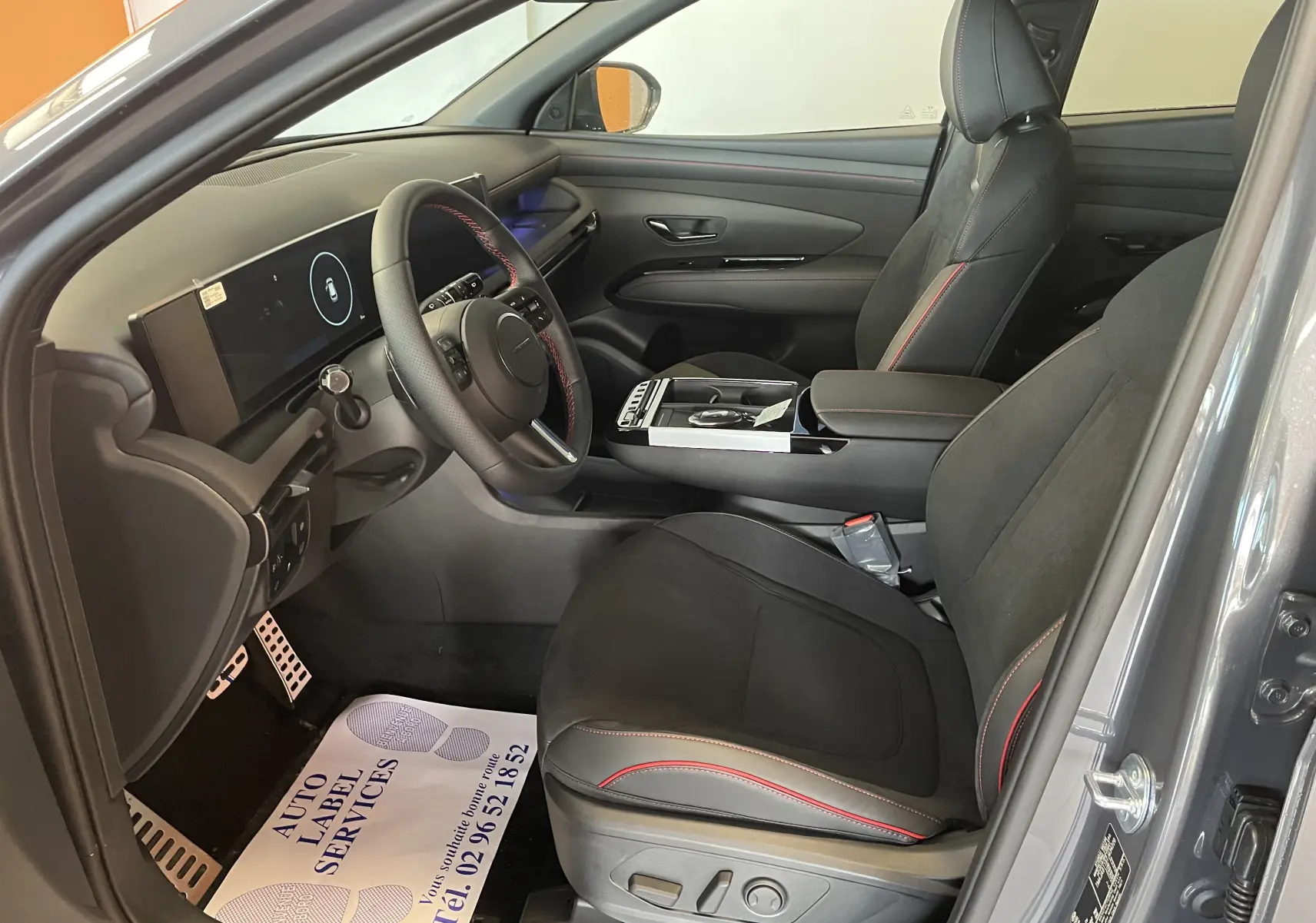 Intérieur du Hyundai Tucson 2025 gris Ecotronic, vue côté gauche, sièges noirs avec surpiqûres rouges et tableau de bord numérique.