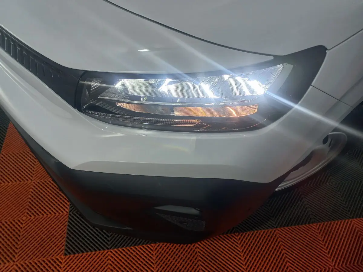 Gros plan sur le phare avant droit allumé d’un Citroën Berlingo blanc, avec pare-chocs noir visible.