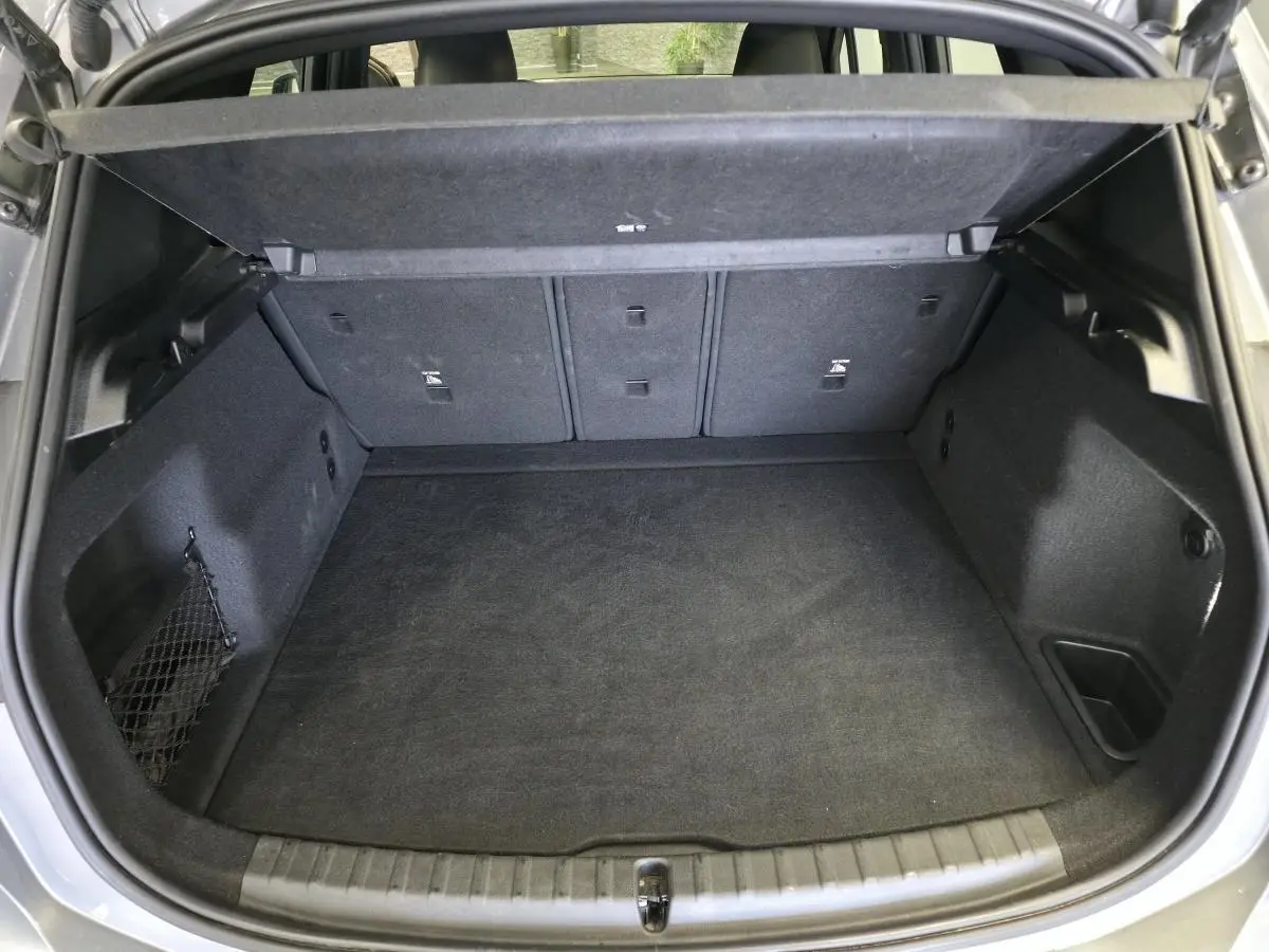 Coffre ouvert vu de l'arrière d'une BMW Série 2 218i Active Tourer gris avec tapis noir et filet de rangement latéral.