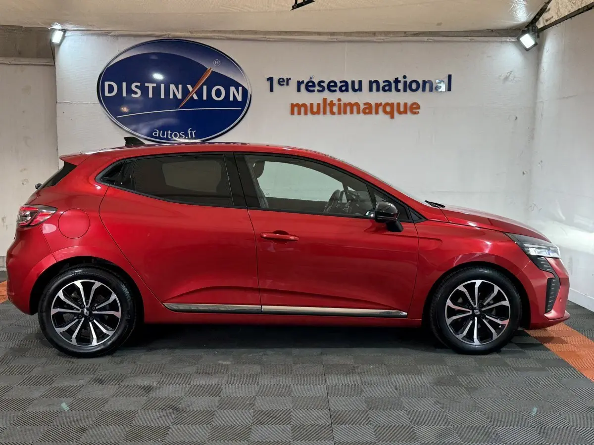 Profil droit d'une Renault Clio Techno TCe 90 rouge 2024, avec jantes alliage et feux LED visibles en intérieur showroom.