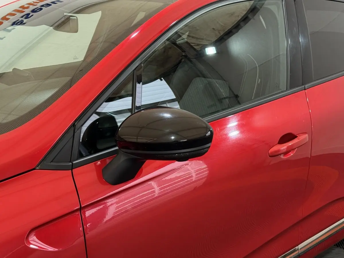 Vue rapprochée du côté gauche d’une Renault Clio Techno TCe 90 rouge, avec rétroviseur noir brillant et poignée de porte assortie.