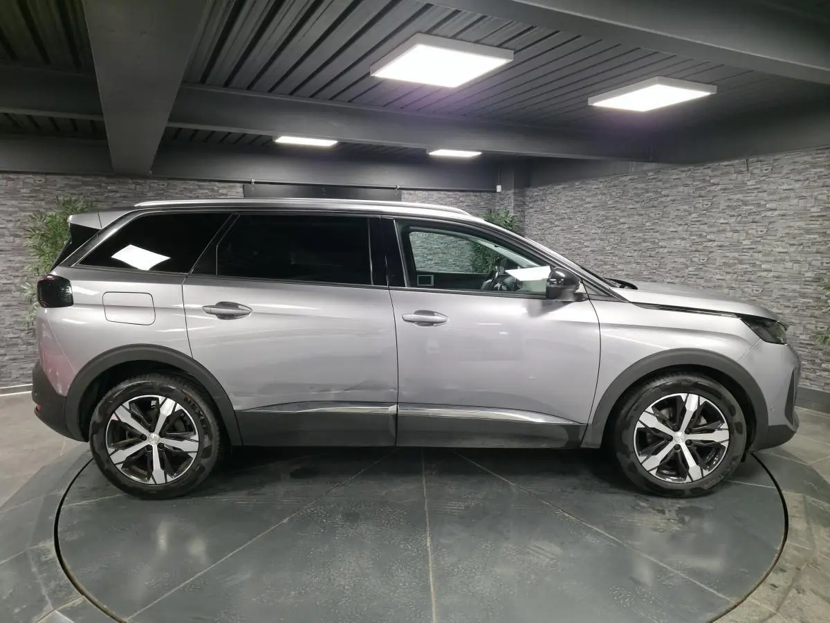 Profil droit du Peugeot 5008 gris métallisé 2022 avec jantes alliage bi-ton et vitres surteintées en intérieur showroom.