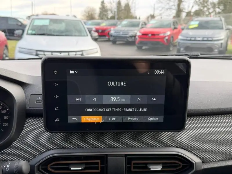 Vue intérieure centrée sur l'écran multimédia du tableau de bord d'une Dacia Sandero blanche, avec affichage radio FM.