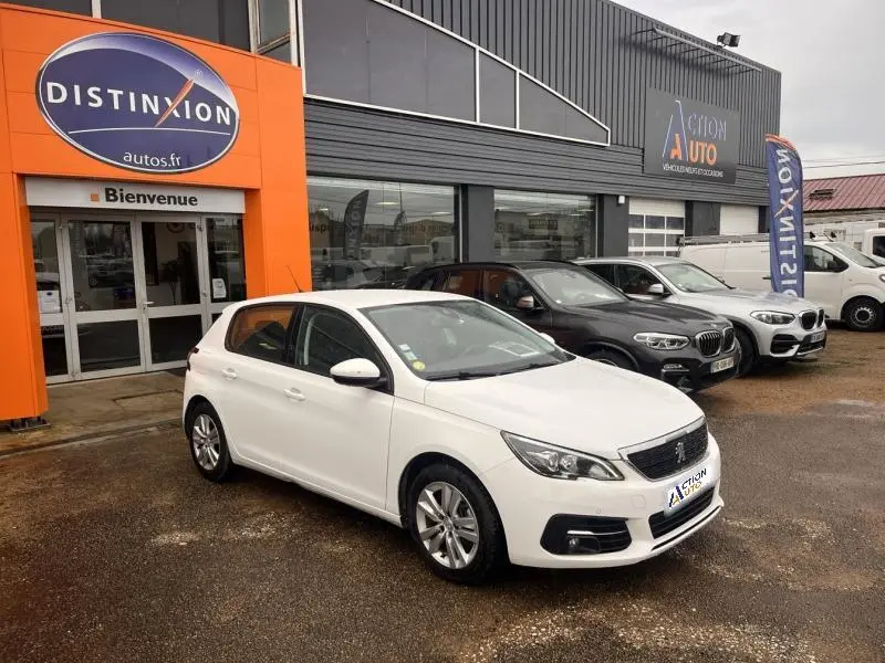 Peugeot 308 blanc banquise vue 3/4 avant droit devant un garage avec plusieurs autres véhicules stationnés.