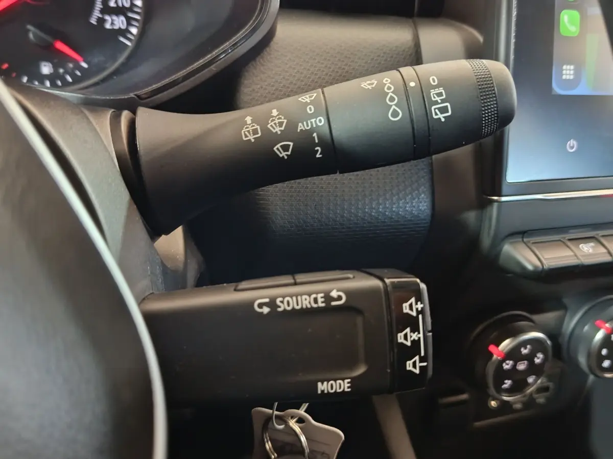 Gros plan sur les commandes au volant et la manette d’essuie-glace d’une Renault Clio gris clair, tableau de bord partiel visible.