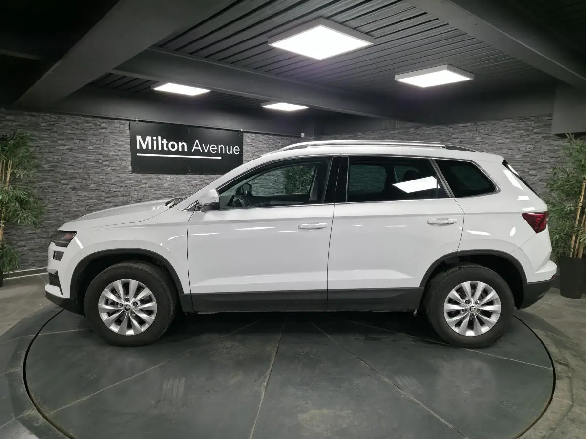 Vue de profil côté gauche d'un SUV Skoda Karoq blanc 2024 avec jantes alliage et barres de toit dans un showroom.