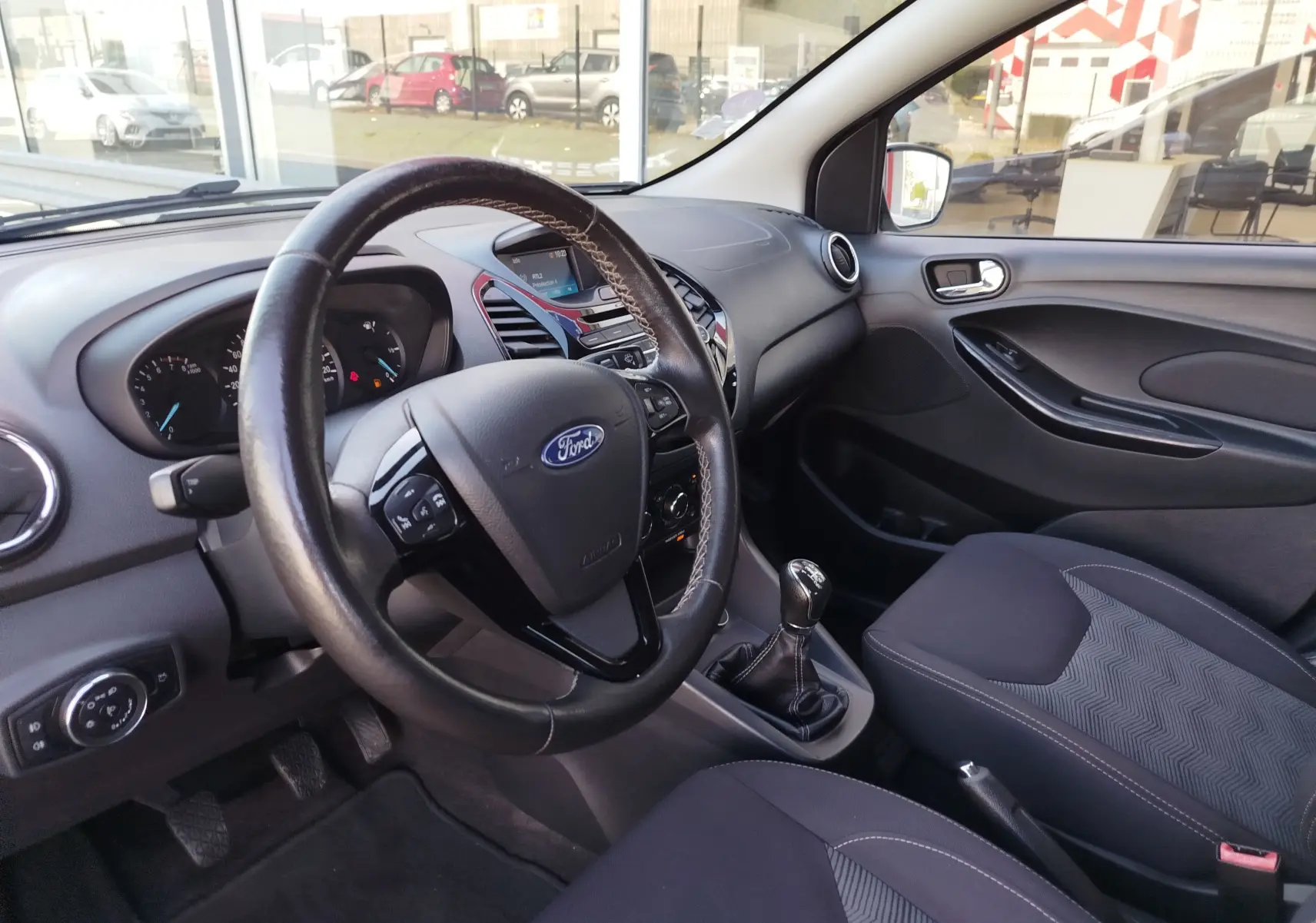 Vue intérieure côté conducteur de la Ford KA+ 2020, volant cuir, tableau de bord noir et sièges tissu gris.