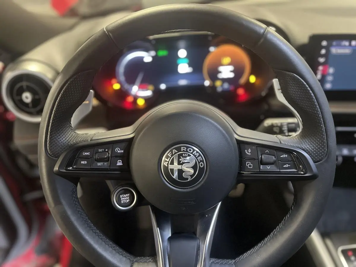 Volant sport en cuir noir avec commandes intégrées et bouton start/stop, tableau de bord digital lumineux en arrière-plan.