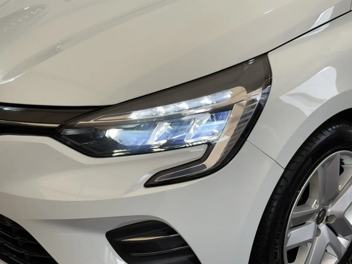 Gros plan sur le phare avant droit allumé d’une Renault Clio Business blanche avec jante alliage visible.