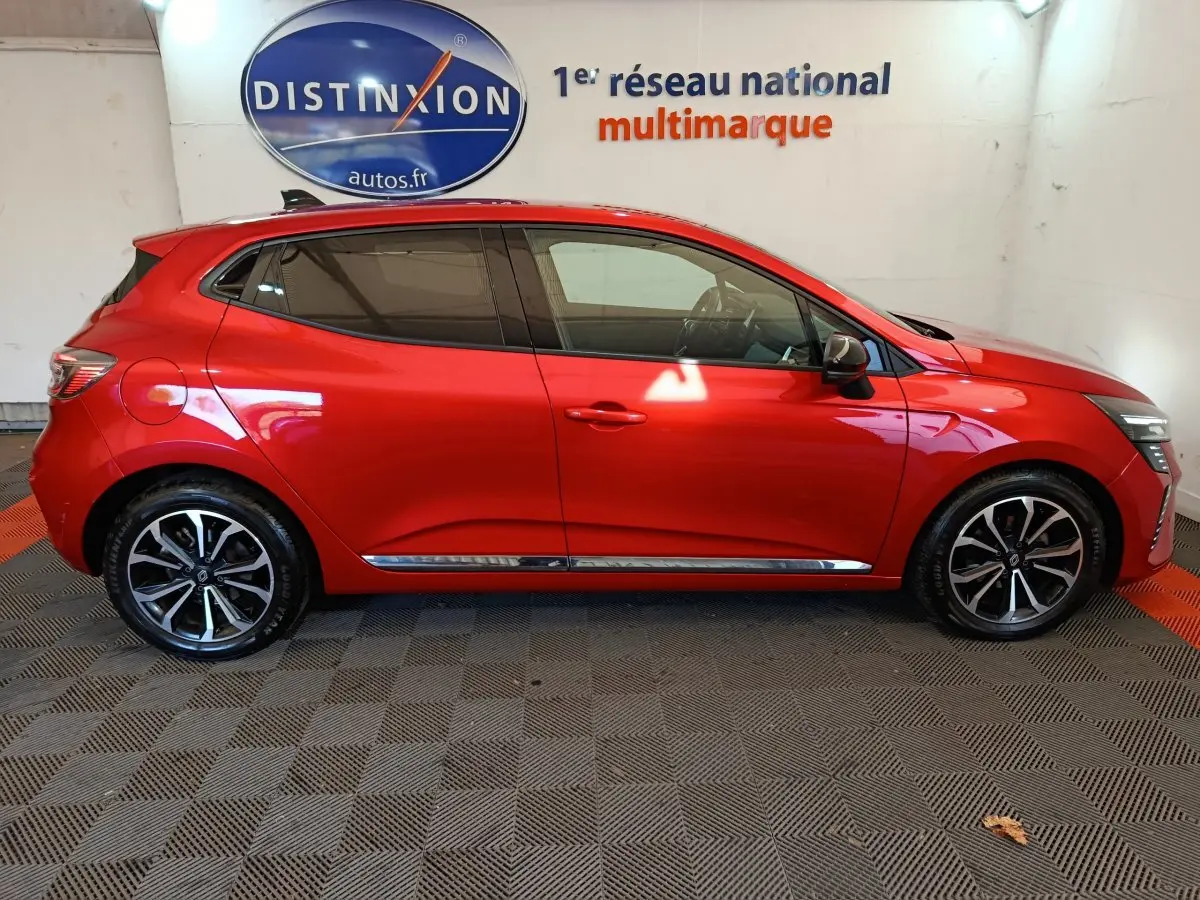 Profil droit d'une Renault Clio Techno TCe 90 rouge 2024 avec jantes alliage et vitres teintées, en intérieur showroom.