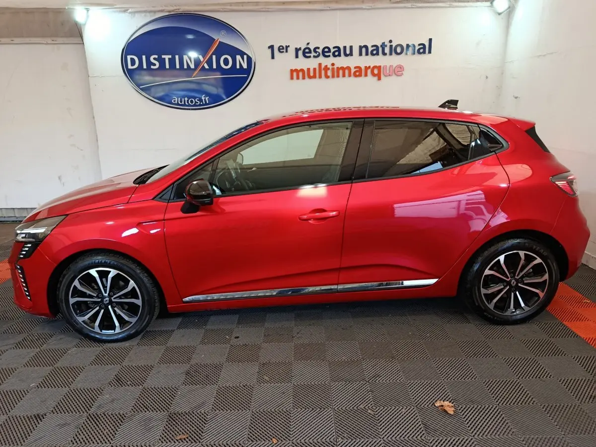 Profil côté gauche d'une Renault Clio Techno TCe 90 rouge 2024, avec jantes alliage et vitres teintées, en intérieur showroom.