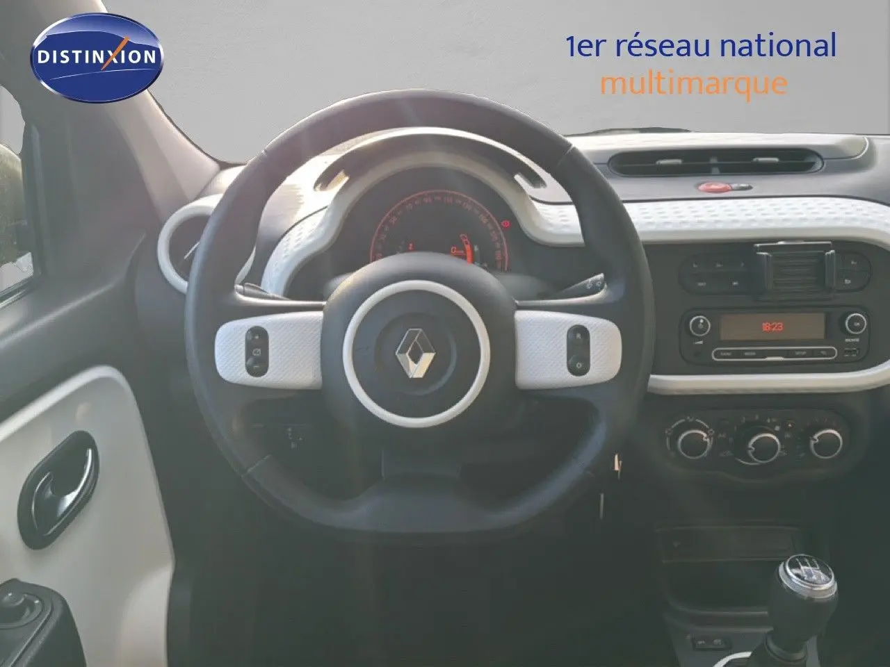 Vue intérieure centrée sur le volant noir et blanc de la Renault Twingo gris acier, avec tableau de bord et levier de vitesses manuelle visibles.