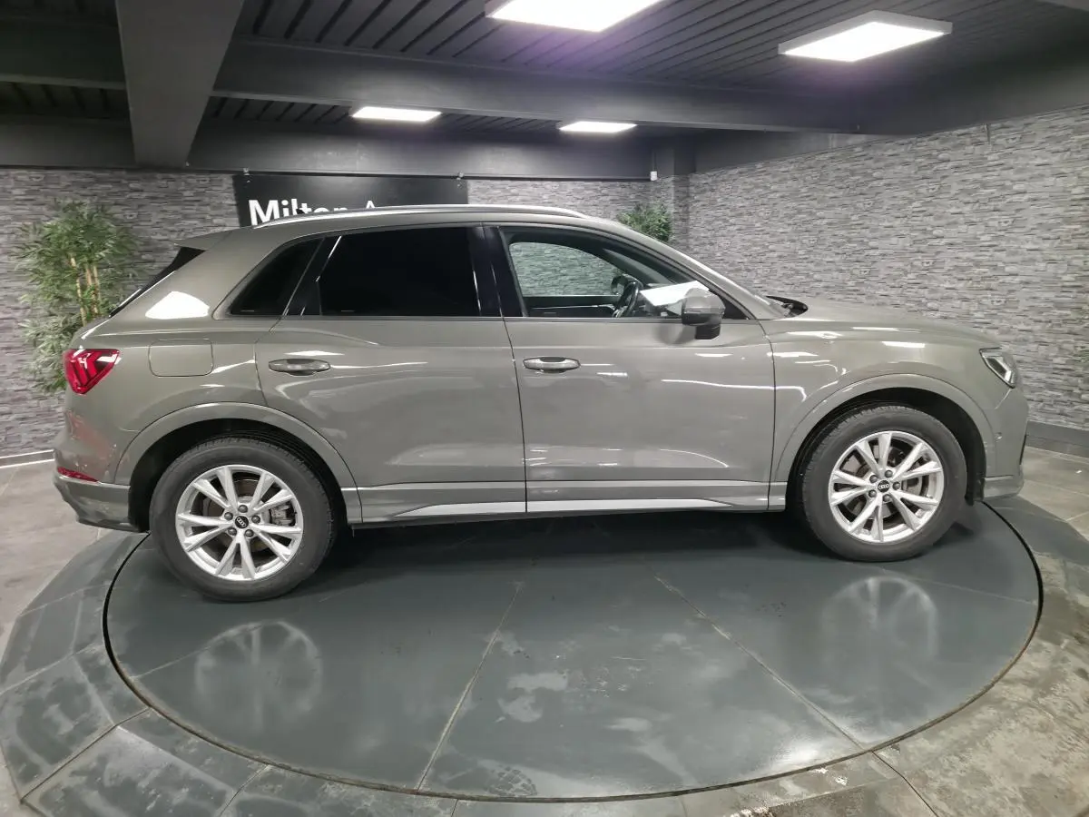 Audi Q3 gris en vue profil côté gauche, vitres surteintées et jantes alliage 5 branches en V visibles en intérieur showroom.