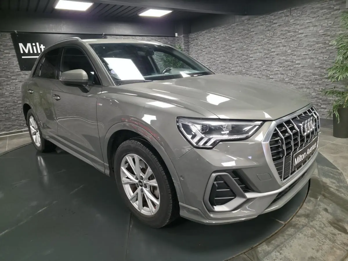 Audi Q3 gris en 3/4 avant droit, avec calandre large et phares LED distinctifs, stationné en intérieur showroom.