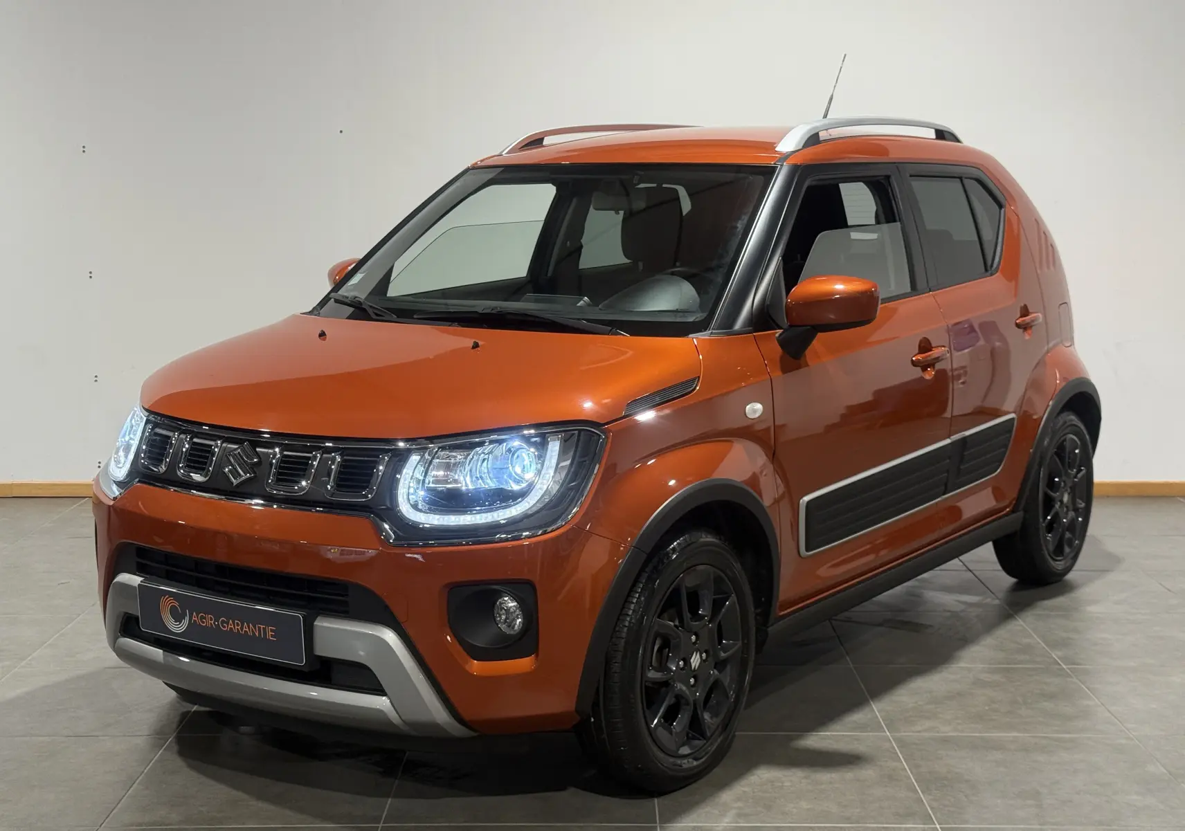 Suzuki Ignis orange vue 3/4 avant droit avec phares allumés et protections latérales noires visibles.