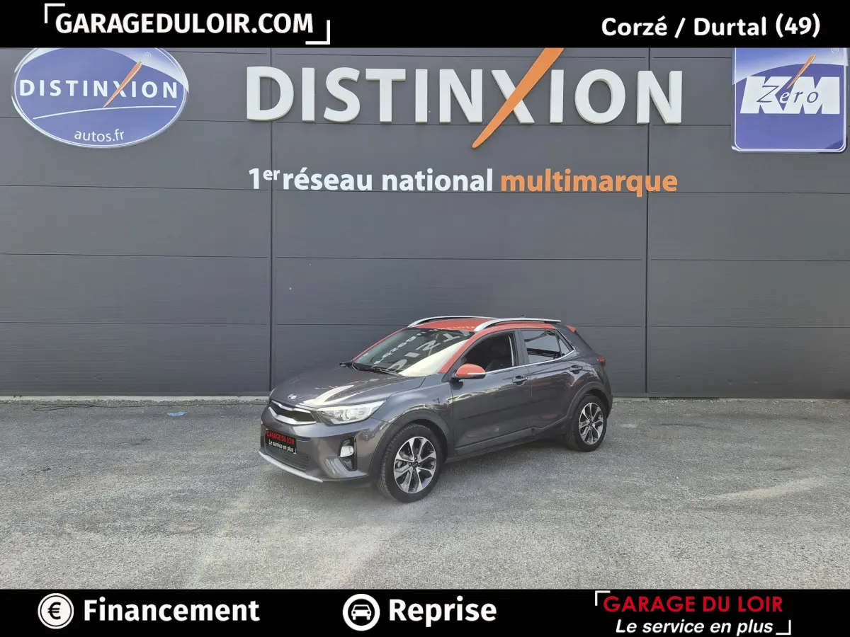 Vue 3/4 avant droite d'un Kia Stonic gris avec toit et rétroviseurs orange devant un mur gris.