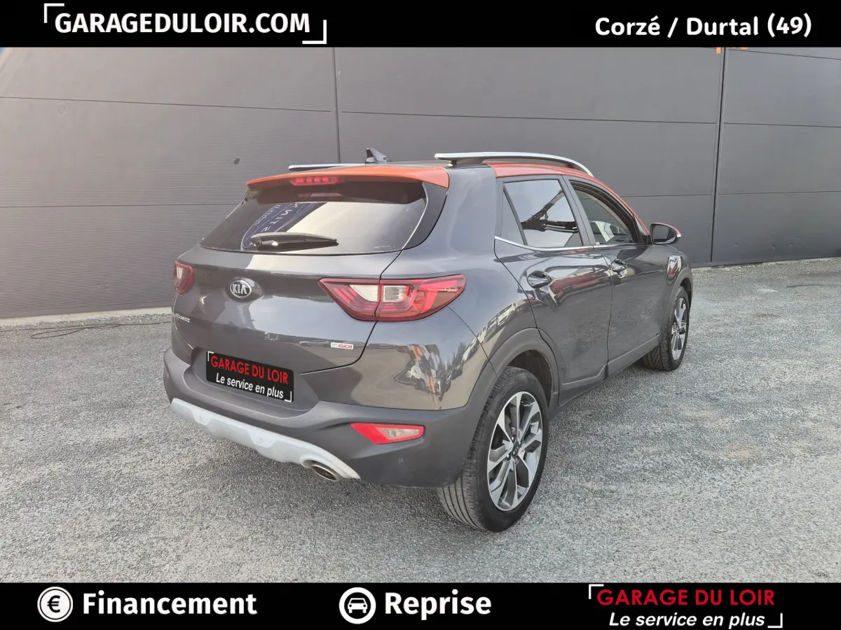 Vue 3/4 arrière droite d’un KIA Stonic 2019 gris foncé avec toit orange et barres de toit chromées.
