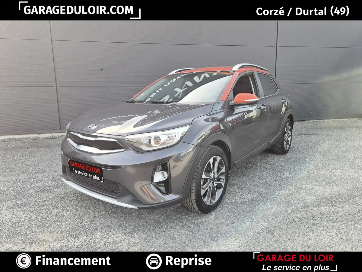 Vue 3/4 avant d'un KIA Stonic gris avec toit et rétroviseurs orange, sur fond urbain neutre.