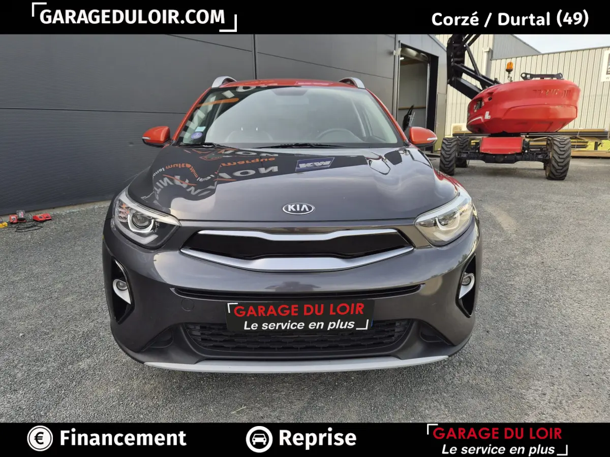 Vue avant d'un KIA Stonic 1.0 T-GDi 100 ACTIVE gris avec rétroviseurs orange, stationné devant un bâtiment industriel.